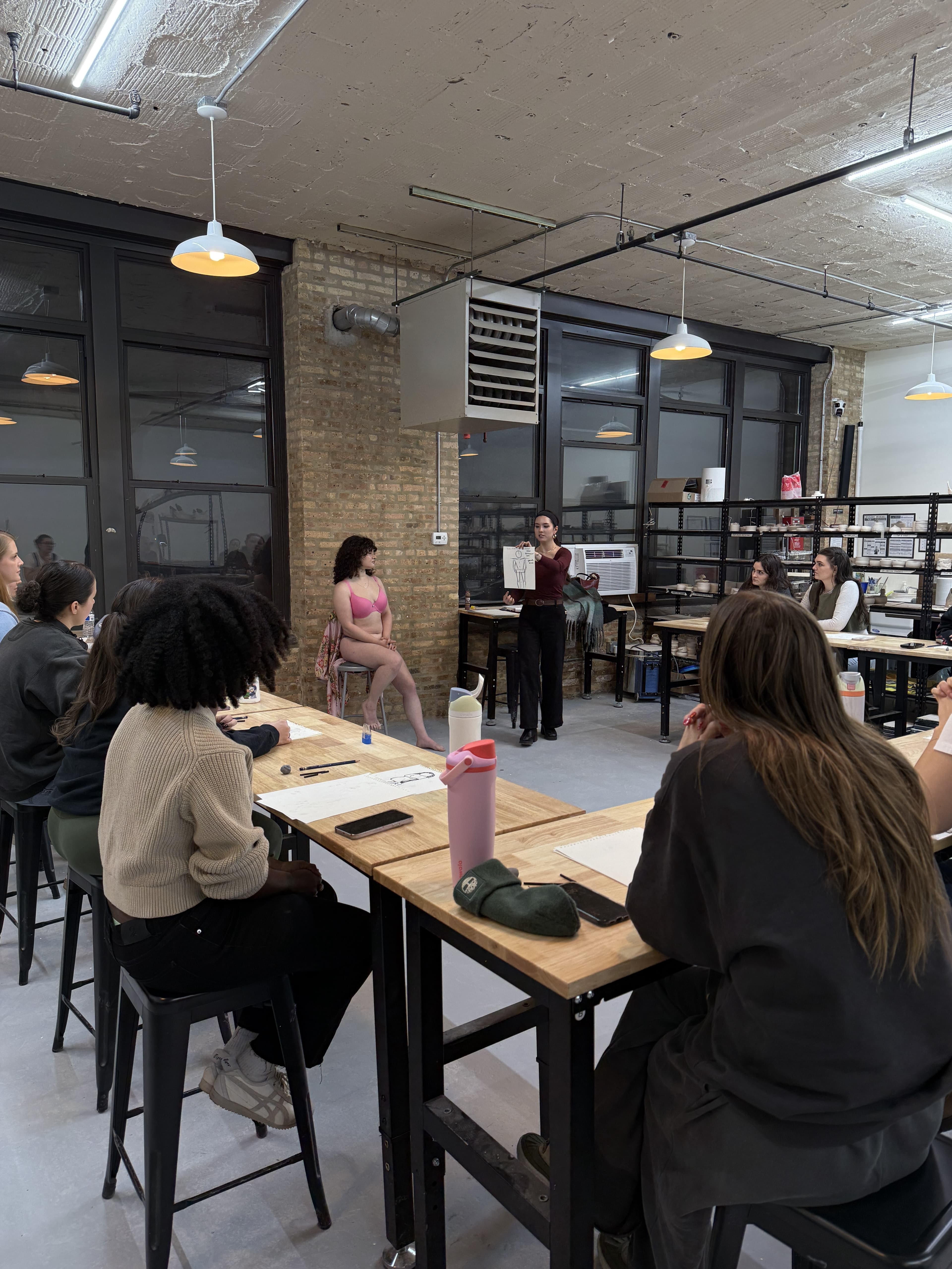 A figure drawing class in session, with the model posing in undergarments and the instructor holding up a demonstration sketch while students draw at shared tables